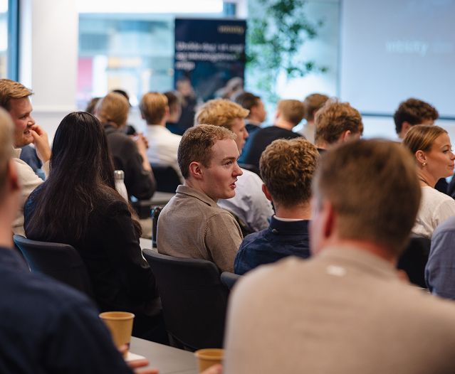 Publikum sitter i rader på en presentasjon eller et seminar innendørs.