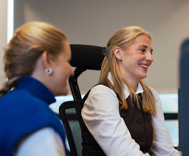  Two women seated in office chairs, looking toward a screen or speaker.