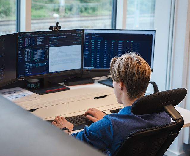 Person working at a desk with three monitors showing code and data.