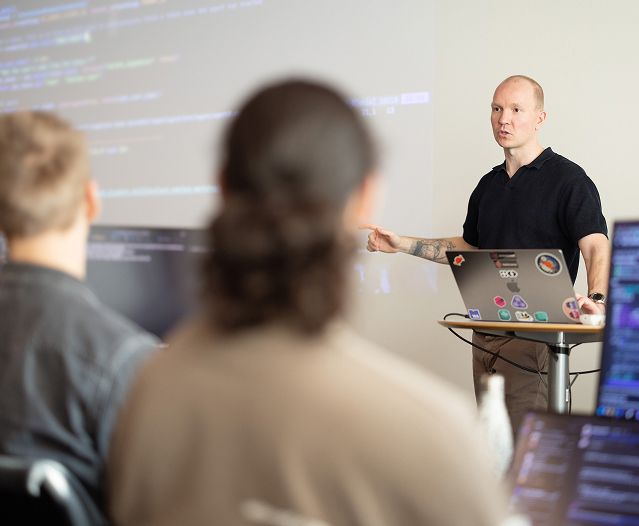 Presenter teaching code to a seated audience with code projected on the wall.