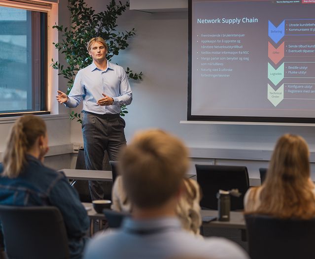 Presenter speaking to an audience beside a slide about network supply chain.