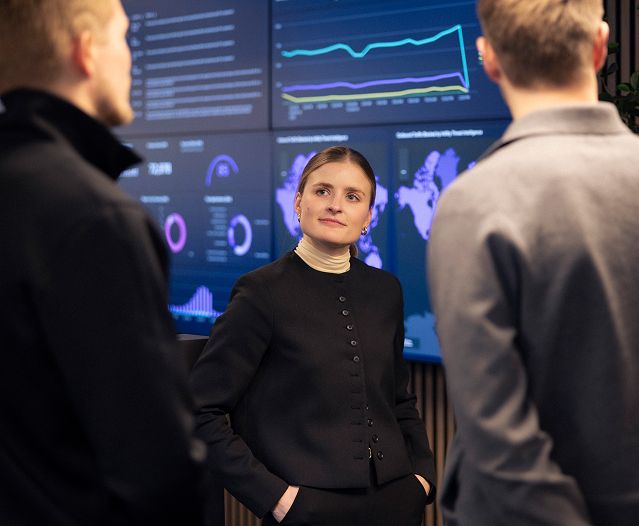 Tre personer diskuterer foran en veggskjerm med grafer og datadashbord.