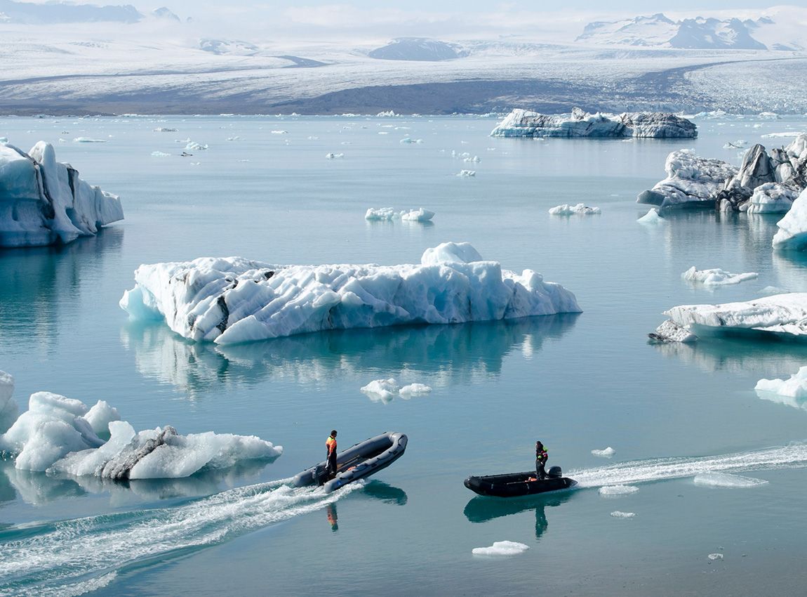 Two people in boats navigate between large icebergs on a calm, icy blue lake surrounded by snowy mountains.
