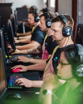 Gamers wearing headphones compete at computers, focusing on screens and keyboards with colorful lights.