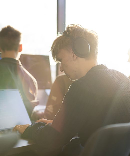Person wearing headphones works on a laptop in a sunlit shared workspace.