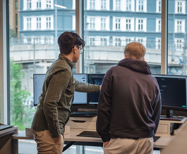 Two people standing at a desk, reviewing code on multiple monitors by a large window.