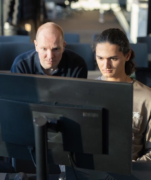 Two colleagues sitting behind a large monitor, working together in a dimly lit office.
