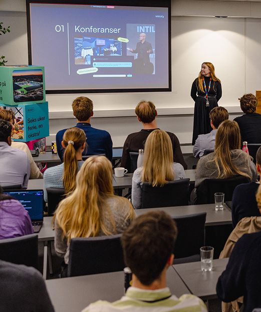 Presenter speaking to an audience in a conference room with a large screen showing conference topics.