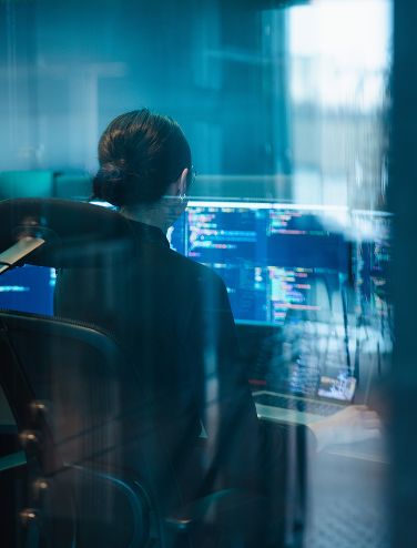 Person in an office chair looking at computer screens with code, seen through glass.