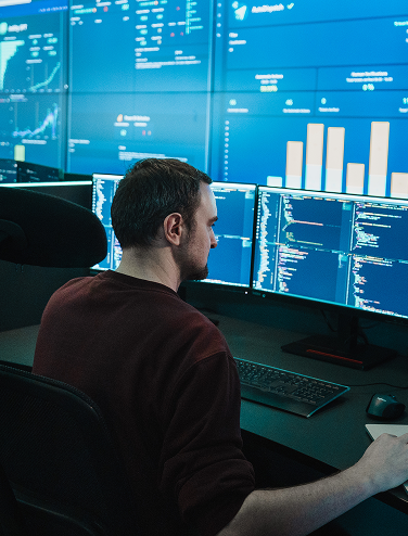 Person working at multiple monitors displaying code and data charts in a modern office.